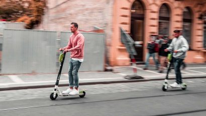 Dos hombres circulando con un patinete eléctrico por la calle.