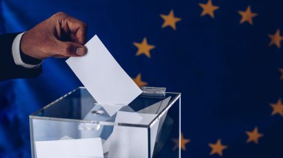 Una persona introduciendo una papeleta dentro de una urna con la bandera europea de fondo.