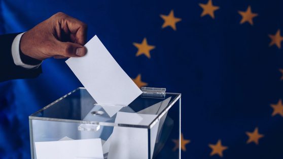Una persona introduciendo una papeleta dentro de una urna con la bandera europea de fondo.